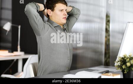 Young businessman and programmer with brown curly hair in a green shirt is stretching his hands after working hard on computer in an office. Business Stock Photo