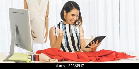 Young Asian Designer woman working and using the technology tablet at workplace over Clothes mannequins, small business startup, Business owner entrep Stock Photo