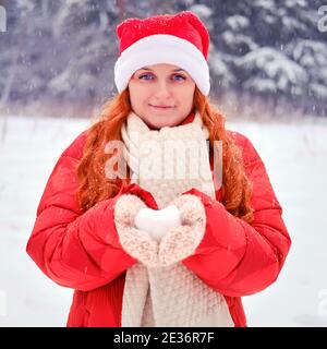 Woman holding snow in the shape of a heart in knitted mittens. Red-haired woman on Christmas and New Year Eve walks in the winter in red clothes Stock Photo