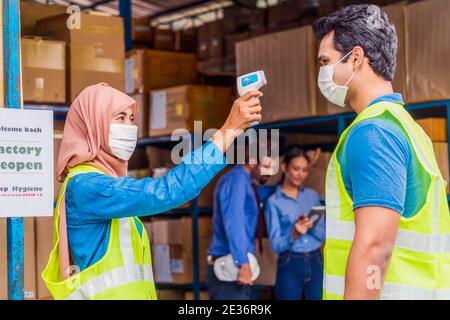 Muslim woman worker wearing surgical mask and Hijab uses Medical Digital Infrared Thermometer measure temperature to Indian man worker with safety clo Stock Photo