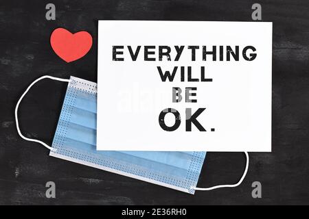 Text 'Everything will be OK' on white paper in front of protective face mask with red heart on dark background Stock Photo