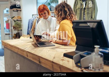 Clothing store workers standing at checkout counter using digital tablet. Two fashion shop partners looking at online orders on a tablet computer in s Stock Photo