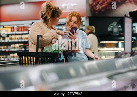 Female shopper showing her cell phone to store assistant and asking for the products at supermarket. Customer at grocery store taking help from a fema Stock Photo