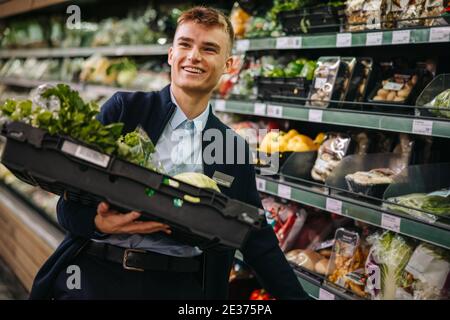 Man working in a supermarket restocking fresh fruit and vegetables in produce section. Young worker working in supermarket. Stock Photo