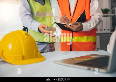 close up helmet of engineer explaining project work on laptop and discussing about drawing blueprint as partner writing data on paper, architect meeti Stock Photo