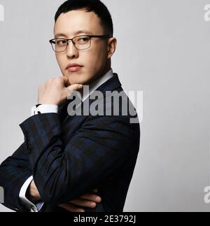 Thoughtful asian businessman expert analyst in formal jacket, tie and glasses stands side to camera with hand at chin Stock Photo