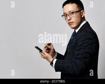 Young confident asian businessman in checkered official suit, tie and glasses standing and making notes in smartphone with stick Stock Photo