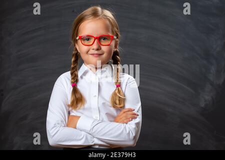 Portrait of a little girl schoolgirl in glasses on the background of a school board. Smart confident child has crossed arms and is looking at the came Stock Photo