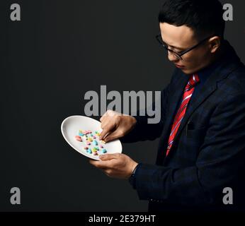 Young asian businessman pharmacist doctor in official suit and glasses holds plate of medical pills taking one Stock Photo