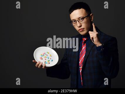 Young asian pharmacist doctor in official suit and glasses holds shows plate of medical pills and holds forefinger up Stock Photo
