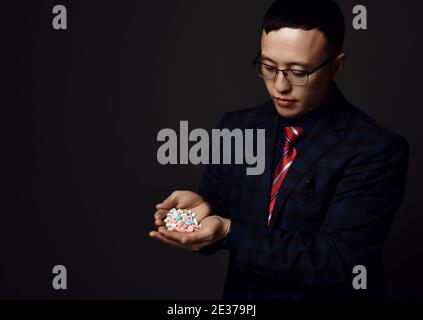 Young asian businessman doctor pharmacist in checkered official suit and glasses holds heap of medical pills Stock Photo