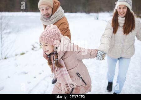 High angle portrait of playful teenage girl holding hands with parents and tugging them along while enjoying family walk in winter forest Stock Photo