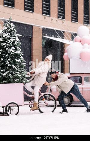 Happy girl sitting on old pink bicycle, boyfriend pushing the bike from behind, having fun in winter Stock Photo
