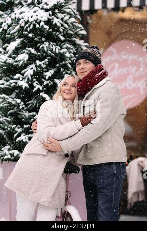 Stylish romantic couple embracing, having fun in winter stand by old pink bicycle during snowfall Stock Photo