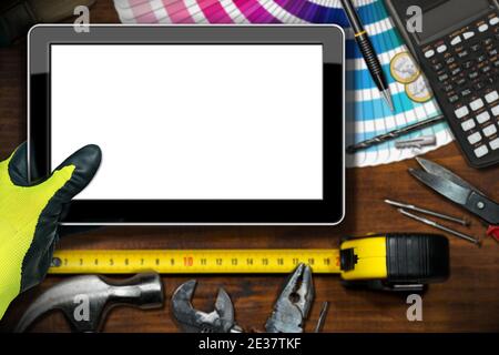 Hand with work glove holding a blank digital tablet on a wooden workbench with DIY work tools, calculator and Euro currency. Stock Photo
