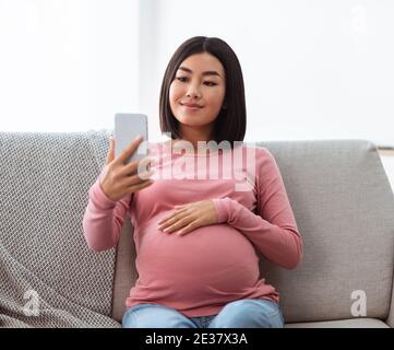 Pregnant Chinese Lady Using Phone Making Selfie Sitting At Home Stock Photo