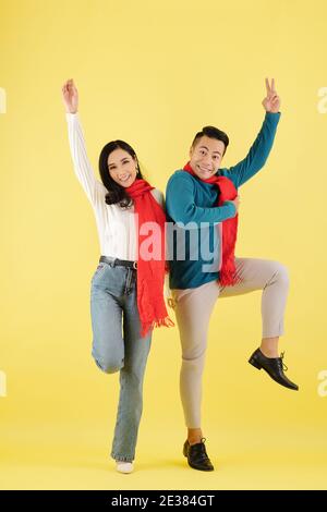 Excited happy young Asian couple in wool winter scarf dancing together against yellow background Stock Photo