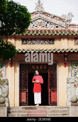Calm pensive young Asian woman in traditional dress standing at open doors of temple with paper fan in hands Stock Photo