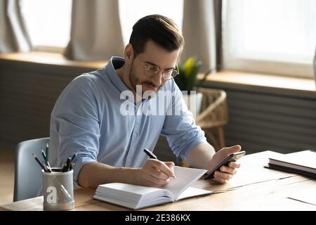 Happy focused young 30s businessman writing notes in paper planner. Stock Photo