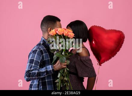 Loving African American couple kissing behind bunch of roses, holding heart shaped balloon on pink studio background Stock Photo