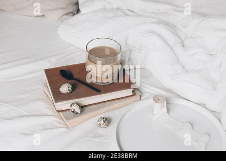 Cozy Easter, spring still life scene. Cup of coffee in glass, books and quail eggs in bedroom. Breakfast in bed concept. Vintage feminine lifestyle Stock Photo