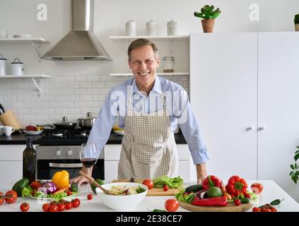 Smiling senior 50s man wearing apron standing in kitchen table preparing salad. Stock Photo