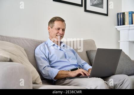 Smiling middle aged man using laptop computer sitting on couch at home. Stock Photo