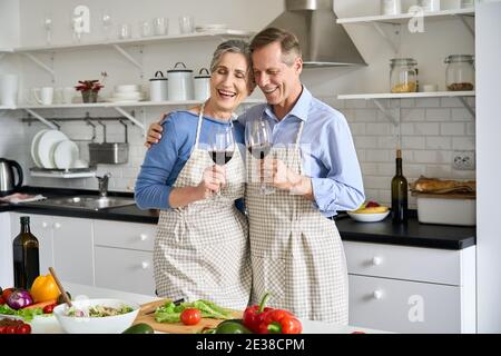 Happy old couple drinking wine preparing vegetable salad in kitchen at home. Stock Photo