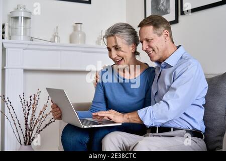 Happy old retired couple using laptop sitting on couch at home together. Stock Photo