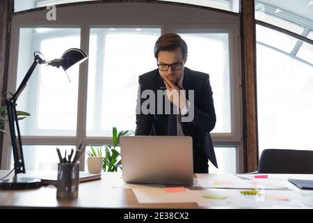 Thoughtful businessman wearing glasses touching chin, using laptop, reading news Stock Photo