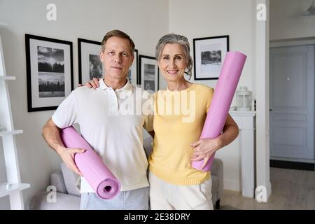 Happy fit old couple hugging, looking at camera, holding yoga mats at home. Stock Photo