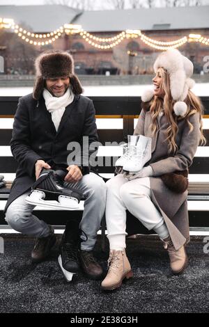 Stylish couple of lovers in warm clothes sitting on bench holding skates at winter ice rink outdoors