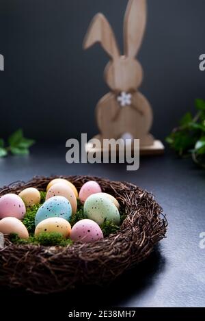 A wicker nest with moss and colored Easter eggs and wooden bunny rabbit and green plant leave on dark background. Happy Easter holiday. Dark mood Stock Photo
