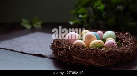A wicker nest with moss and colored Easter eggs on gray linen fabric napkin and few green leaves on dark background. Dark mood style. Happy Easter Stock Photo