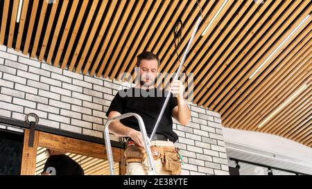 A male electrician carries out a service for installing LED lamps in a designer loft-style wooden ceiling. Rent-a-gent carries out work on replacement Stock Photo