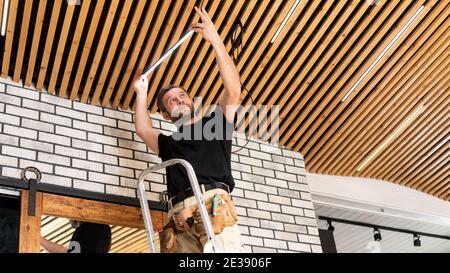 An electrician installs an LED lamp on a wooden plank ceiling. Repair of household electrical networks. Lamp replacement service. Installation of a mo Stock Photo