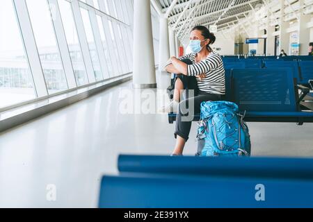 Lonely female solo traveler with backpack sitting in the empty airport passenger transfer hall in protective face mask and looking out large windows. Stock Photo