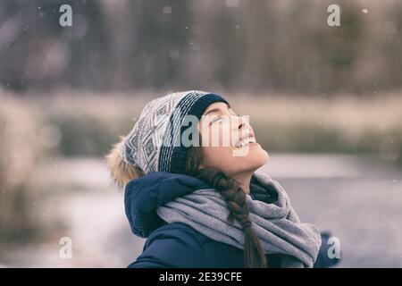 Winter woman happy enjoying snow falling on face outdoor forest nature Asian girl looking up wearing hat and scarf cold weather clothes Stock Photo