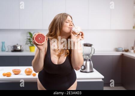 Choosing healthy or unhealthy food positive woman standing on the kitchen holding grapefruit and daughnut with lots fruits on table next to blander Stock Photo
