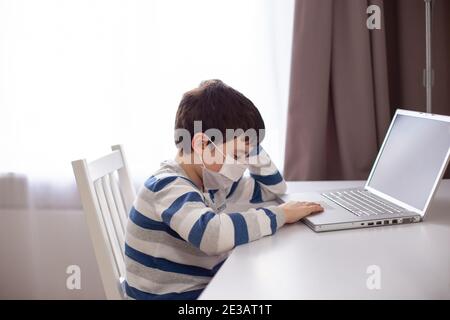 A boy in a white medical mask , sits behind a laptop