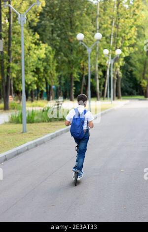 a handsome stylish and young schoolboy in a white shirt, blue tie and a backpack with a scooter on a street. back view Stock Photo
