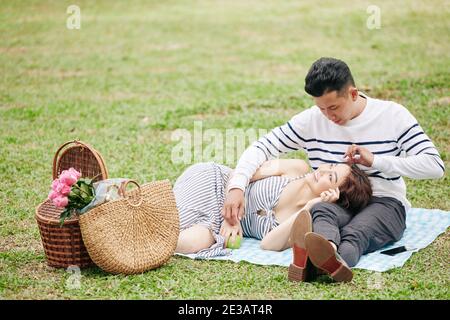 Young Vietnamese man in love looking at girlfriend resting head on his laps when they are having picnic in park Stock Photo