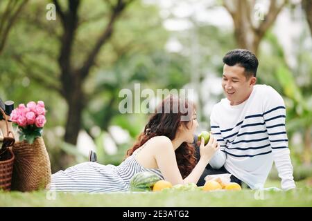 Smiling man offering girlfriend a bite of green apple when they are having picnic Stock Photo