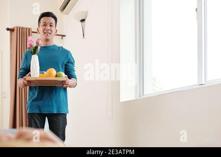 Caring young Asian man bringing tray with Valentines day breakfast and flowers in bedroom Stock Photo
