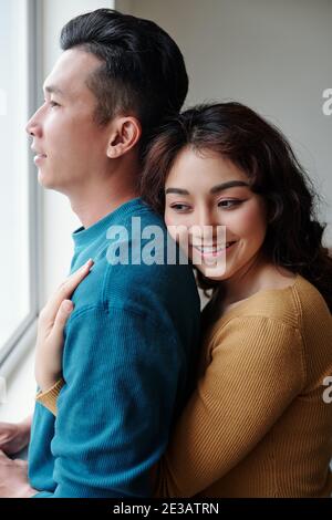 Happy pretty young woman hugging her husband from behind when they are standing near window Stock Photo