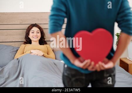 Young man hiding romantic Valentines day present for his girlfriend snuggling in bed after waking up Stock Photo