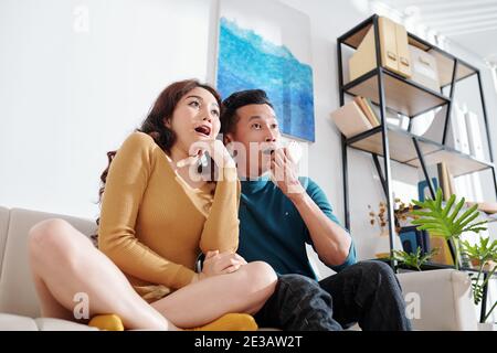 Frightened young Asian couple watching scary movie at home Stock Photo