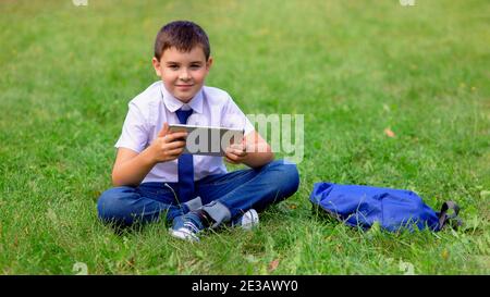 A happy schoolboy in a white shirt sits on green grass and hold a tablet Stock Photo