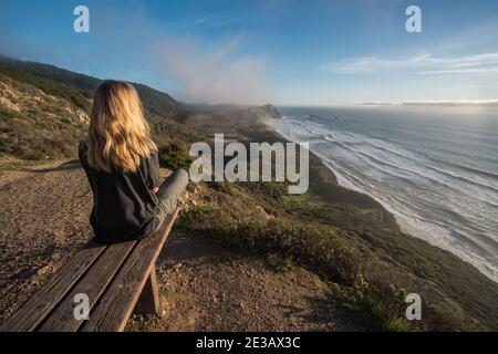A blonde hiker rests on a bench that overlooks the pacific ocean in a beautiful portion of the California coastline in Point Reyes national seashore.