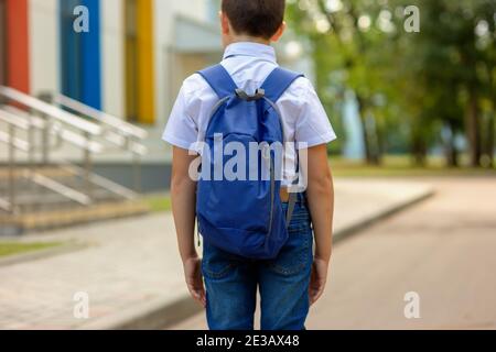 A brunette schoolboy in a white shirt and blue backpack goes to school Stock Photo